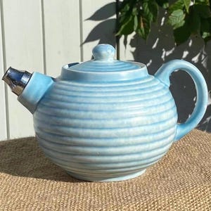 May include: A light blue ceramic teapot with a horizontal ribbed texture, a matching lid with a small knob, a curved handle, and a silver-toned metal spout attachment. The glaze features subtle variations in color intensity across the surface.