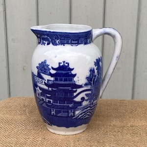May include: A white ceramic pitcher featuring a blue transferware pattern. The design depicts a detailed scene with a multi-story pagoda, stylized trees, and architectural elements. The pitcher has a curved handle and a pouring spout at the top.