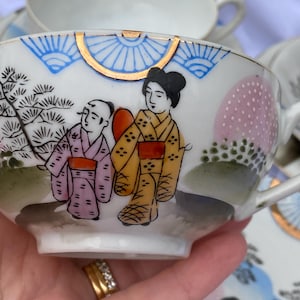Puede incluir: Taza de té de porcelana blanca con una escena japonesa pintada a mano. La taza presenta un borde dorado, detalles azules y dorados, y una representación de dos figuras con vestimenta tradicional. Elementos de paisaje rosas y verdes completan el diseño.
