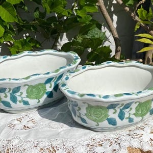 May include: Two white ceramic planters with scalloped edges. Each planter is decorated with green rose and blue leaf designs. The planters are oval-shaped and sit on a white lace tablecloth. The background features green foliage.