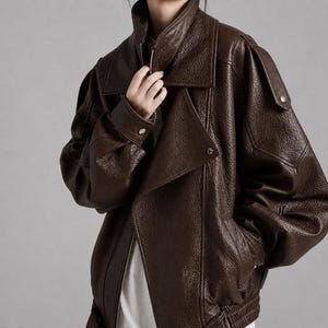 Peut inclure: Une veste en cuir marron foncé avec une finition texturée et grainée, dotée d'un large col, de manches longues avec poignets boutonnés, de pattes d'épaule et d'une fermeture éclair frontale. La veste a une coupe ample avec un ourlet côtelé.