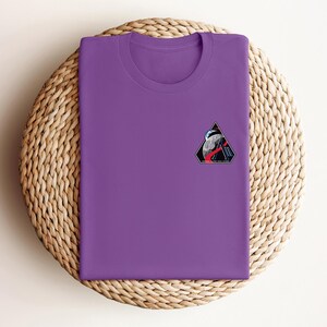 May include: A purple crew neck t-shirt with a small, colorful patch on the chest. The patch features a triangular design with a space-themed illustration, including a planet, astronaut, and geometric shapes. The t-shirt is folded neatly on a woven, circular surface.