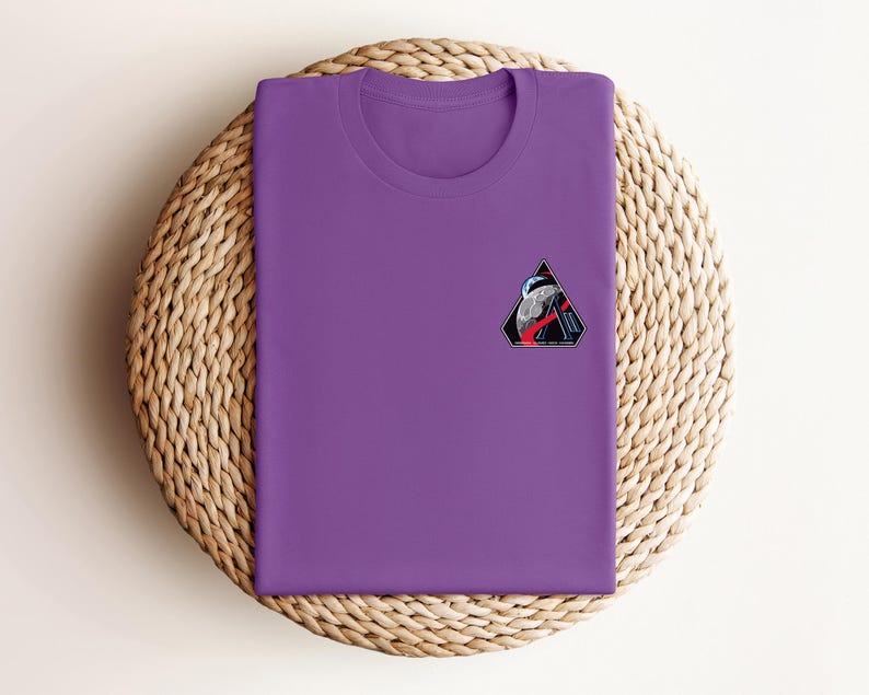 May include: A purple crew neck t-shirt with a small, colorful patch on the chest. The patch features a triangular design with a space-themed illustration, including a planet, astronaut, and geometric shapes. The t-shirt is folded neatly on a woven, circular surface.