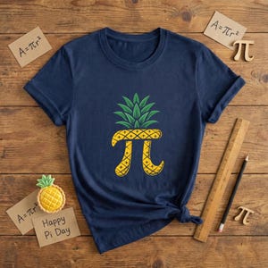 May include: Navy blue t-shirt featuring a pineapple design with a pi symbol. The pineapple has green leaves and a yellow body with a crosshatch pattern. The shirt is on a wooden surface with "Happy Pi Day" text.