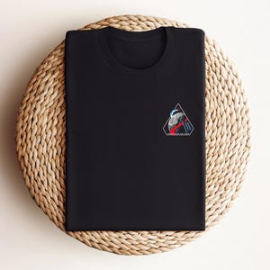 May include: Black crew neck t-shirt with a small, colorful graphic on the chest. The graphic is a geometric design with red, white, and blue elements. The shirt is folded neatly on a woven, circular surface.