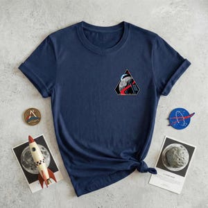 May include: Navy blue t-shirt with a small, triangular patch featuring a space-themed design. The patch includes a planet, a rocket, and the text "NASA". A small, round Artemis pin and a rocket model are also visible.