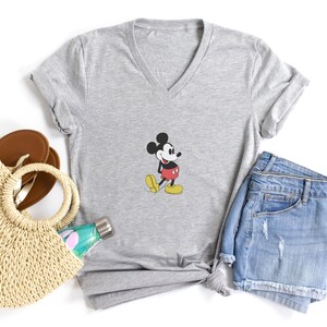Puede incluir: Una camiseta gris con cuello en V con un gr&aacute;fico de Mickey Mouse. La imagen tambi&eacute;n incluye pantalones cortos de mezclilla, un bolso de mano de paja, sandalias marrones y una botella de agua, todo dispuesto sobre una superficie blanca. La camiseta tiene un dise&ntilde;o de dibujos animados cl&aacute;sico.