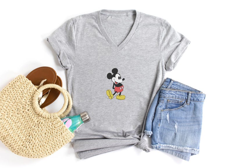 Puede incluir: Una camiseta gris con cuello en V con un gr&aacute;fico de Mickey Mouse. La imagen tambi&eacute;n incluye pantalones cortos de mezclilla, un bolso de mano de paja, sandalias marrones y una botella de agua, todo dispuesto sobre una superficie blanca. La camiseta tiene un dise&ntilde;o de dibujos animados cl&aacute;sico.