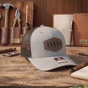 May include: Gray and black trucker hat with a leather patch that reads "DAD EST. 2018". The hat is on a wooden surface with tools, pens, and a notebook in the background.