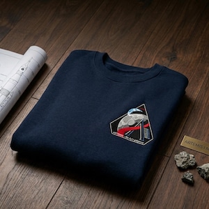 May include: Navy blue sweatshirt with an embroidered Artemis I mission patch. The patch features a stylized moon, rocket, and the text "Artemis I Mission". A blueprint and a small metal plaque are also visible.