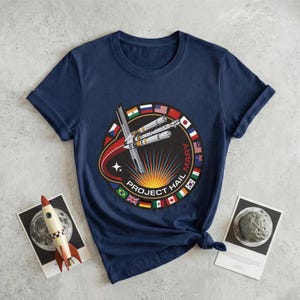 May include: Navy blue t-shirt featuring a circular graphic with a rocket, flags, and the text "PROJECT HAIL MARY". The design includes a red and orange comet trail and a star. The shirt is knotted at the bottom.