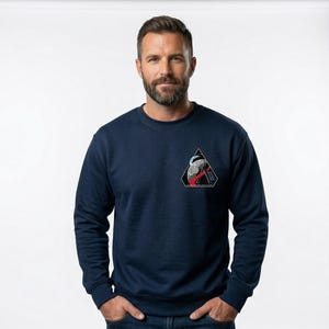 May include: Navy blue sweatshirt with a triangular patch featuring a graphic design in red, white, and black. The sweatshirt has long sleeves and a crew neck. The person in the image is wearing blue jeans.