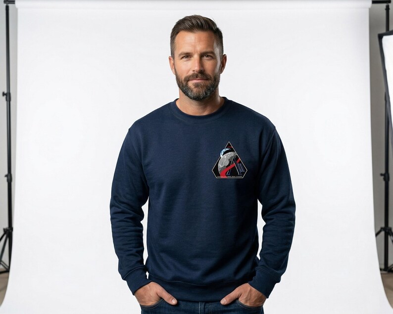 May include: Navy blue sweatshirt with a triangular patch featuring a graphic design in red, white, and black. The sweatshirt has long sleeves and a crew neck. The person in the image is wearing blue jeans.