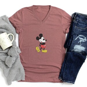 Puede incluir: Una camiseta color malva con cuello en V con un gr&aacute;fico cl&aacute;sico de Mickey Mouse, combinada con jeans azules desgastados, una bufanda gris y una taza blanca. El personaje de dibujos animados est&aacute; en su pose ic&oacute;nica, con pantalones cortos rojos y zapatos amarillos.