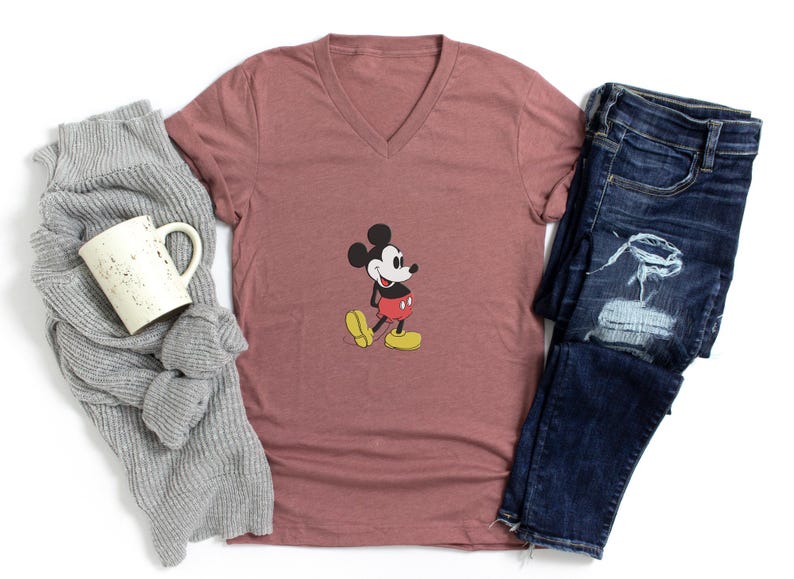 Puede incluir: Una camiseta color malva con cuello en V con un gr&aacute;fico cl&aacute;sico de Mickey Mouse, combinada con jeans azules desgastados, una bufanda gris y una taza blanca. El personaje de dibujos animados est&aacute; en su pose ic&oacute;nica, con pantalones cortos rojos y zapatos amarillos.