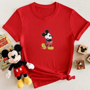 Puede incluir: Una camiseta roja con un dise&ntilde;o de Mickey Mouse. La imagen tambi&eacute;n incluye un peluche de Mickey Mouse, un c&oacute;mic vintage de Mickey Mouse y bloques de madera con dise&ntilde;os de Mickey Mouse.