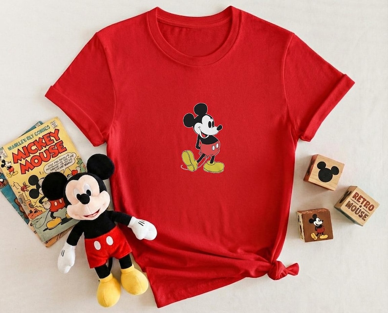 Puede incluir: Una camiseta roja con un dise&ntilde;o de Mickey Mouse. La imagen tambi&eacute;n incluye un peluche de Mickey Mouse, un c&oacute;mic vintage de Mickey Mouse y bloques de madera con dise&ntilde;os de Mickey Mouse.