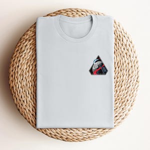 May include: A light gray t-shirt with a small, colorful patch featuring a moon, a rocket, and a triangular design. The shirt is folded neatly on a woven, circular placemat.