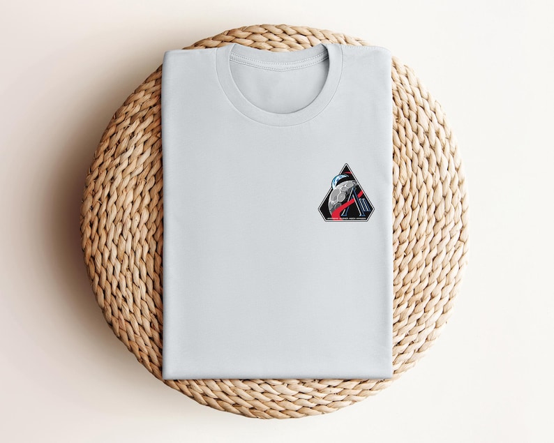 May include: A light gray t-shirt with a small, colorful patch featuring a moon, a rocket, and a triangular design. The shirt is folded neatly on a woven, circular placemat.