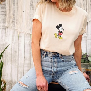 Puede incluir: Camiseta color crema con un gr&aacute;fico de Mickey Mouse. El personaje de dibujos animados est&aacute; representado en negro, rojo, amarillo y blanco. La camiseta se combina con vaqueros de mezclilla azul claro con detalles desgastados.