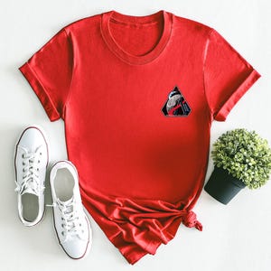 May include: A red t-shirt with a small graphic patch on the chest, paired with white canvas sneakers. The graphic patch is a black triangle with a design inside. A small potted plant is also in the image.
