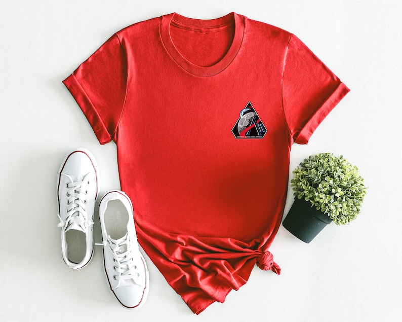 May include: A red t-shirt with a small graphic patch on the chest, paired with white canvas sneakers. The graphic patch is a black triangle with a design inside. A small potted plant is also in the image.