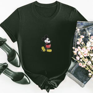 Puede incluir: Camiseta negra con un peque&ntilde;o dise&ntilde;o bordado de Mickey Mouse. La camiseta se combina con pantalones cortos de mezclilla, tacones negros y un ramo de flores rosas. La camiseta tiene un cuello redondo cl&aacute;sico y mangas cortas.
