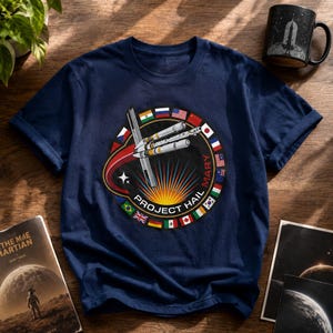 May include: A navy blue t-shirt featuring a circular graphic with the text Project Hail Mary. The design includes a central spacecraft illustration, a red curved trail, a bright yellow sunburst, and a border of various international flags.