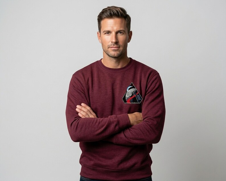 May include: Maroon crew neck sweatshirt with a triangular patch featuring a graphic design. The patch includes a black triangle with a white and red design. The sweatshirt is a solid color and has long sleeves.