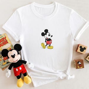 Puede incluir: Camiseta blanca con un dibujo de Mickey Mouse. El personaje de dibujos animados est&aacute; representado con pantalones cortos rojos, zapatos amarillos y guantes negros. Un peluche de Mickey Mouse y un c&oacute;mic vintage tambi&eacute;n est&aacute;n en la imagen.