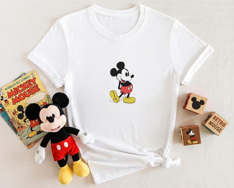 Puede incluir: Camiseta blanca con un dibujo de Mickey Mouse. El personaje de dibujos animados est&aacute; representado con pantalones cortos rojos, zapatos amarillos y guantes negros. Un peluche de Mickey Mouse y un c&oacute;mic vintage tambi&eacute;n est&aacute;n en la imagen.