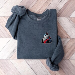 May include: A dark gray sweatshirt with a crew neck and long sleeves. A triangular patch on the chest features a space-themed design with a planet, astronaut, and the text "SPACE FORCE ACADEMY". The sweatshirt is folded on a wooden surface.