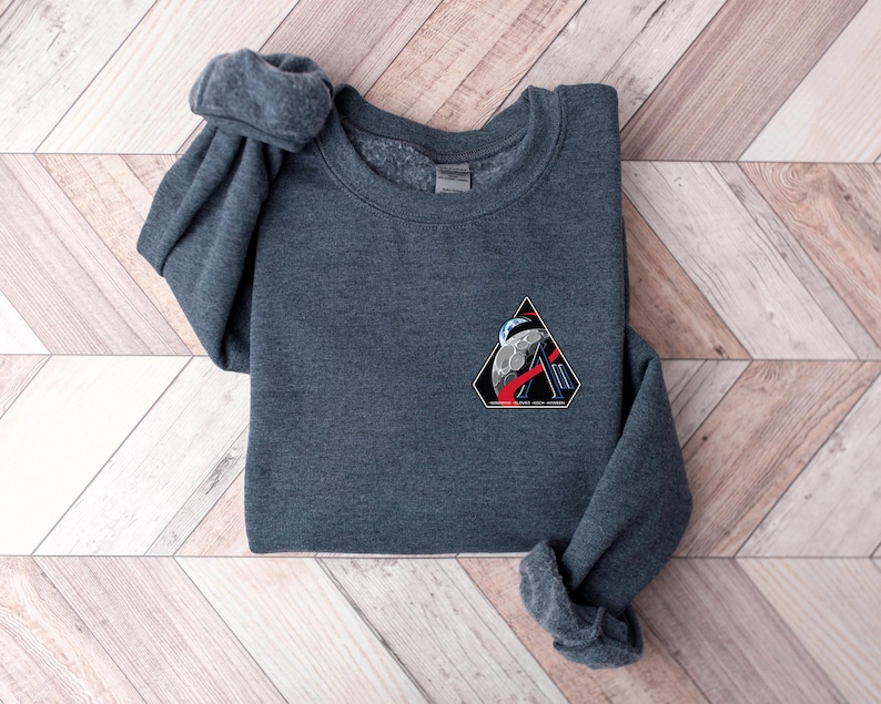 May include: A dark gray sweatshirt with a crew neck and long sleeves. A triangular patch on the chest features a space-themed design with a planet, astronaut, and the text "SPACE FORCE ACADEMY". The sweatshirt is folded on a wooden surface.