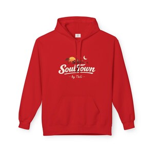 May include: A red hoodie with a front pocket. The design features the words "Soul Town" in a stylized font, with a graphic of a sunset, buildings, and a crescent moon. The text "by T&G" is below the main text.