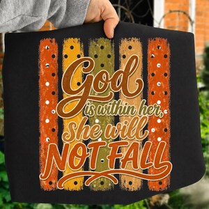 Peut inclure: Un t-shirt noir avec un motif graphique composé de cinq bandes verticales texturées dans des tons orange, jaune et vert. Le texte indique God is within her, she will not FALL dans une police marron et blanche avec des petits points blancs.