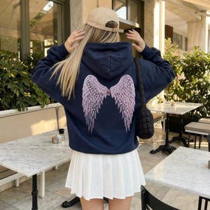 K&ouml;nnte beinhalten: Marineblauer Kapuzenpullover mit hellrosa Engelsfl&uuml;gel-Design auf dem R&uuml;cken. Dazu ein wei&szlig;er Faltenrock und eine beige Baseballkappe. Eine schwarze, gesteppte Umh&auml;ngetasche ist ebenfalls zu sehen.