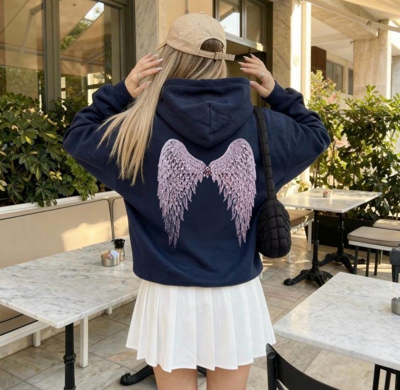 K&ouml;nnte beinhalten: Marineblauer Kapuzenpullover mit hellrosa Engelsfl&uuml;gel-Design auf dem R&uuml;cken. Dazu ein wei&szlig;er Faltenrock und eine beige Baseballkappe. Eine schwarze, gesteppte Umh&auml;ngetasche ist ebenfalls zu sehen.