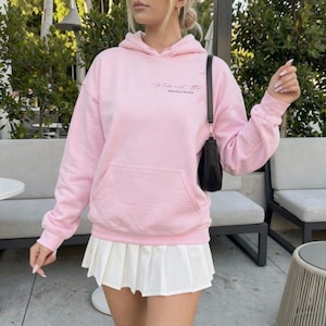 K&ouml;nnte beinhalten: Hellrosa Kapuzenpullover mit Fronttasche und dem Text "The higher you rise, the more they hate". Dazu ein wei&szlig;er Faltenrock und eine schwarze Handtasche.