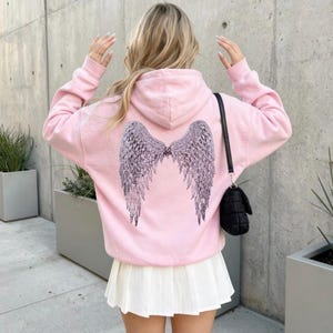 K&ouml;nnte beinhalten: Hellrosa Kapuzenpullover mit einem gro&szlig;en, detaillierten Fl&uuml;geldesign auf dem R&uuml;cken. Die Fl&uuml;gel sind dunkelgrau und haben aufwendige Federdetails. Der Hoodie hat eine Kapuze und lange &Auml;rmel. Die Person tr&auml;gt einen wei&szlig;en Faltenrock und eine schwarze Handtasche.