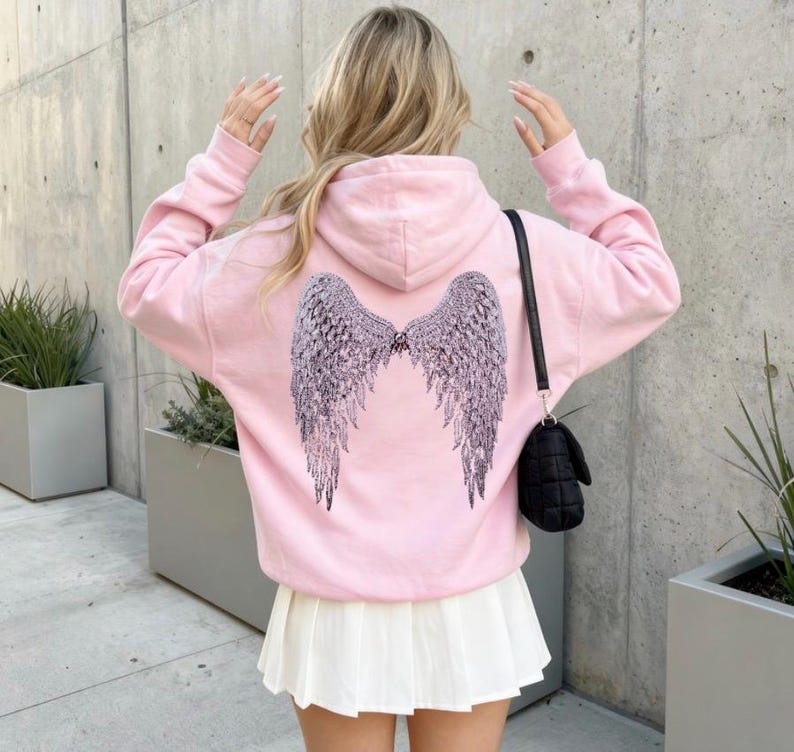 K&ouml;nnte beinhalten: Hellrosa Kapuzenpullover mit einem gro&szlig;en, detaillierten Fl&uuml;geldesign auf dem R&uuml;cken. Die Fl&uuml;gel sind dunkelgrau und haben aufwendige Federdetails. Der Hoodie hat eine Kapuze und lange &Auml;rmel. Die Person tr&auml;gt einen wei&szlig;en Faltenrock und eine schwarze Handtasche.