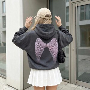 K&ouml;nnte beinhalten: Ein dunkelgrauer Kapuzenpullover mit einem hellvioletten Engelsfl&uuml;gel-Design auf dem R&uuml;cken. Die Person tr&auml;gt einen wei&szlig;en Faltenrock und eine beige Baseballkappe. Eine schwarze Umh&auml;ngetasche ist ebenfalls zu sehen.