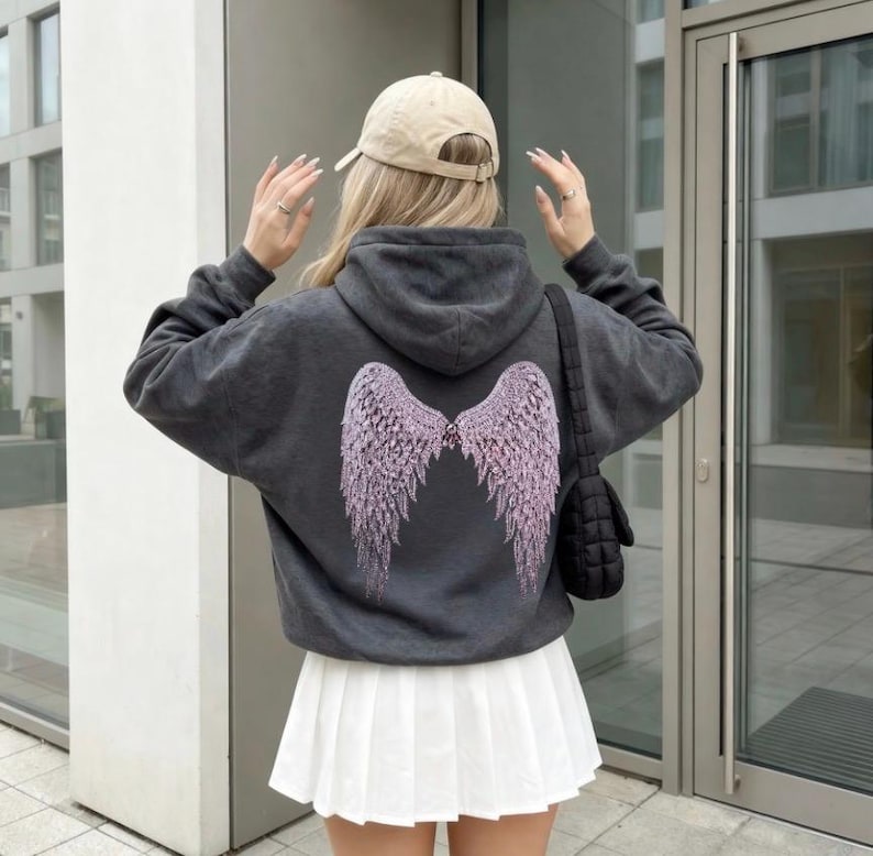 K&ouml;nnte beinhalten: Ein dunkelgrauer Kapuzenpullover mit einem hellvioletten Engelsfl&uuml;gel-Design auf dem R&uuml;cken. Die Person tr&auml;gt einen wei&szlig;en Faltenrock und eine beige Baseballkappe. Eine schwarze Umh&auml;ngetasche ist ebenfalls zu sehen.