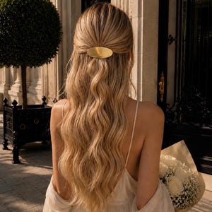 May include: A smooth, oval-shaped gold hair clip secured at the back of long, wavy blonde hair. The hair accessory features a polished, metallic finish and a simple, rounded design that holds a section of hair in a half-up, half-down style.