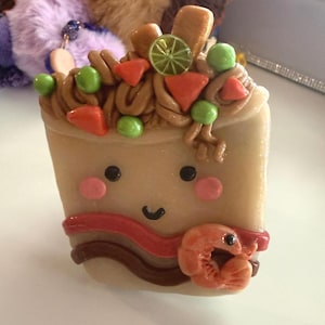 May include: A whimsical, handmade food-themed charm. The charm is a tan-colored square with a smiling face, pink cheeks, and black eyes. It is topped with brown noodles, green peas, orange triangles, and a lime slice. A shrimp is attached to the side.