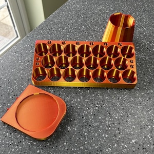 May include: A 3D-printed orange and gold object, possibly a container or organizer, with multiple cylindrical holes. The object is rectangular with a detached lid and a separate, cylindrical piece. The design features a gradient of orange and gold colours.