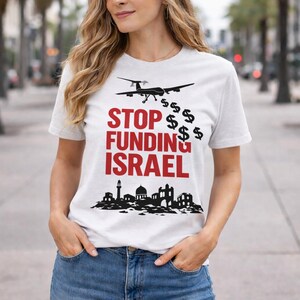 Op de afbeelding: Wit t-shirt met de tekst "STOP FUNDING ISRAEL" in het rood, vergezeld van een drone en dollartekens. De onderkant van het shirt heeft een zwart silhouet van gebouwen.