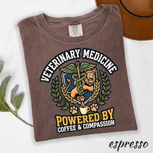 May include: Espresso-colored t-shirt with a graphic design for veterinary medicine. The design includes a lion, a caduceus, and the text "VETERINARY MEDICINE POWERED BY COFFEE & COMPASSION". The shirt is a Comfort Colors brand.
