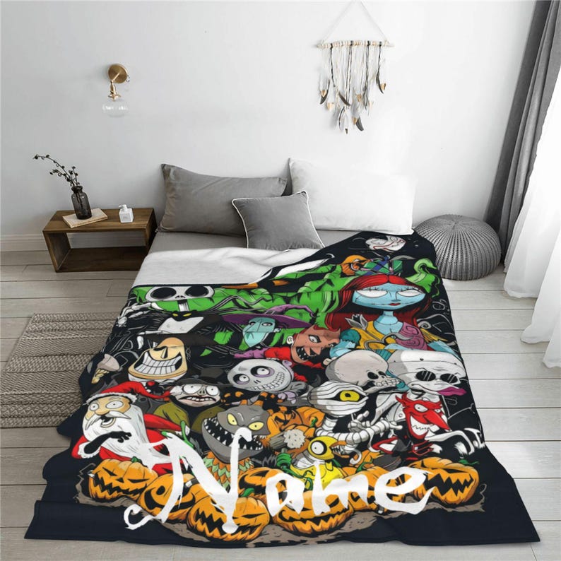 Op de afbeelding: Een zwarte deken met een kleurrijk cartoonontwerp met verschillende personages en pompoenen. Het woord "Name" staat in het wit onderaan. De deken ligt op een bed met grijze kussens en een wit dekbed.