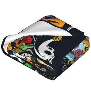 Op de afbeelding: Gevouwen zwarte deken met een kleurrijk cartoonontwerp met een spook, een pompoen en andere personages. De deken heeft een witte binnenkant en een gedetailleerde, levendige buitenkant.