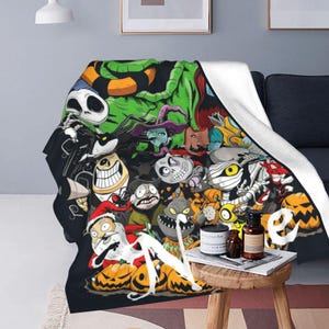 Op de afbeelding: Een zwarte deken met een collage van cartoonfiguren met een Halloween-thema. De deken, met een witte achterkant, is over een donkergrijze bank gedrapeerd. Personages zijn onder meer Jack Skellington, Sally en andere spookachtige figuren.